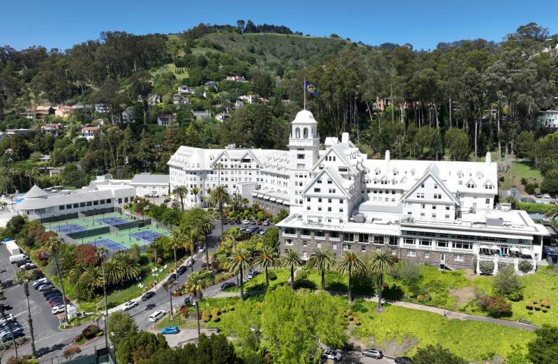 Aerial image of a large, bright white hotel set against a green, tree-covered hillside. The hotel has a castle-like appearance, and there are tennis courts to the left of it.