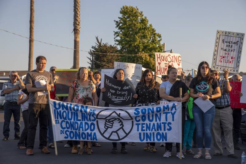 A group of people stand together holding signs. The white sign in the middle reads "Sonoma County Tenants Union.: