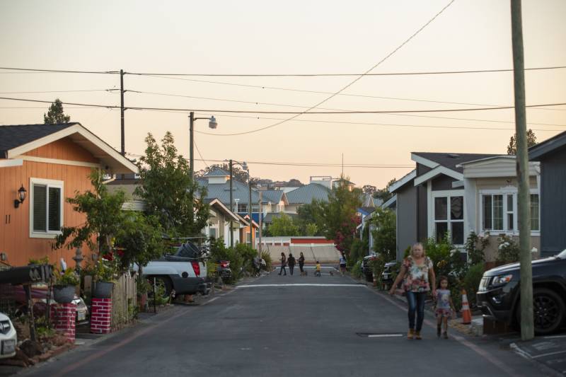 Adults and children are seen on a street running between rows of mobile homes.
