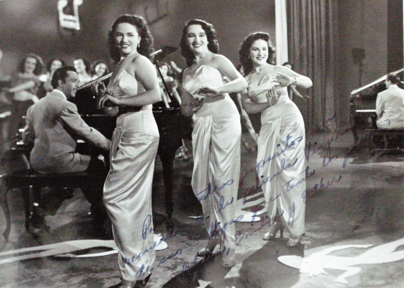 A vintage, black and white photo of three women wearing dresses with a man playing piano in the background.