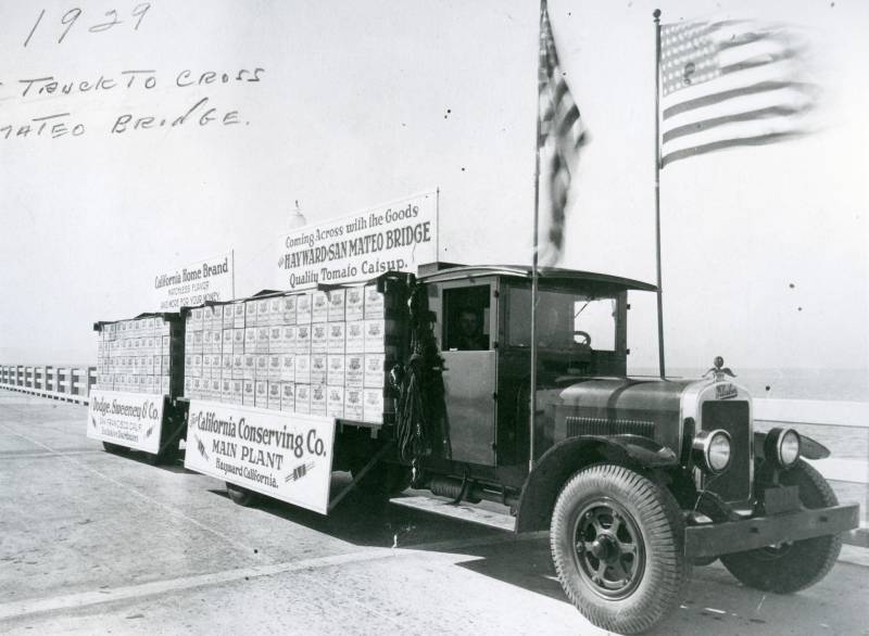 Archival image of a 1920s-era transport truck carrying crates of ketchup. Two large American flags are mounted on poles near the front of the truck.