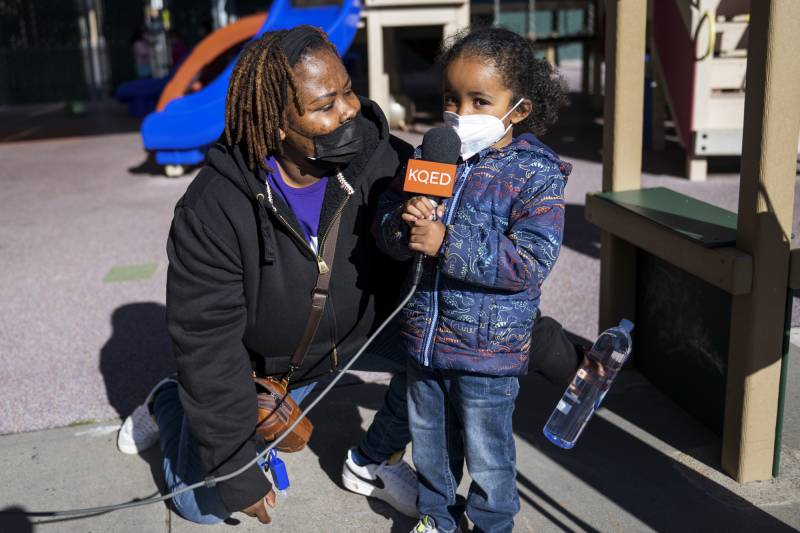 A young person wearing a face mask holds a microphone with KQED branding on it as an adult kneels next to them and looks on.