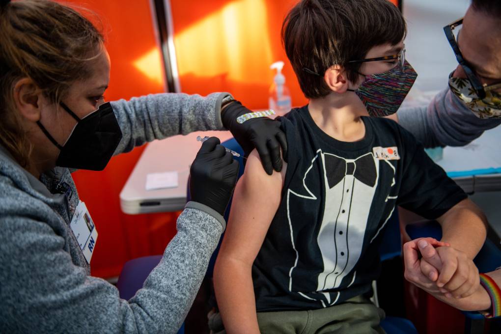 A boy with a tuxedo T-shirt and a face mask holds his mother's hand as he receives a shot in the arm by a nurse.