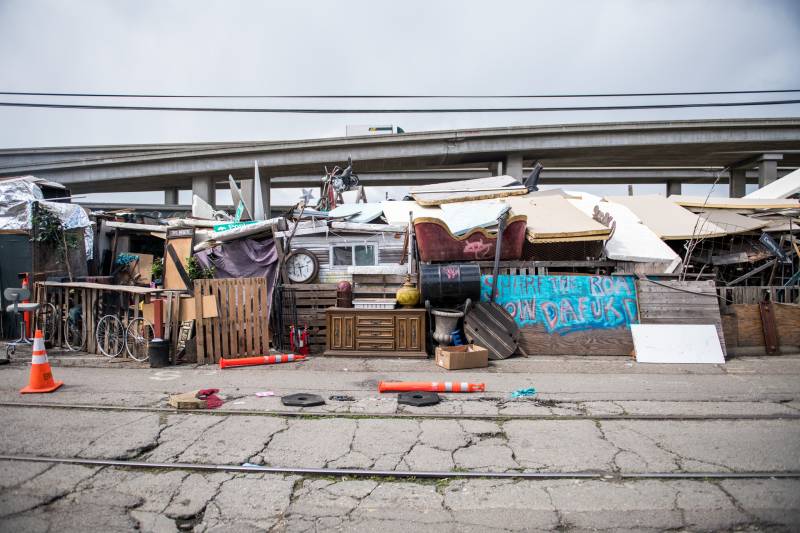 a row of tents and wood boards up against motor homes under a freeway overpass