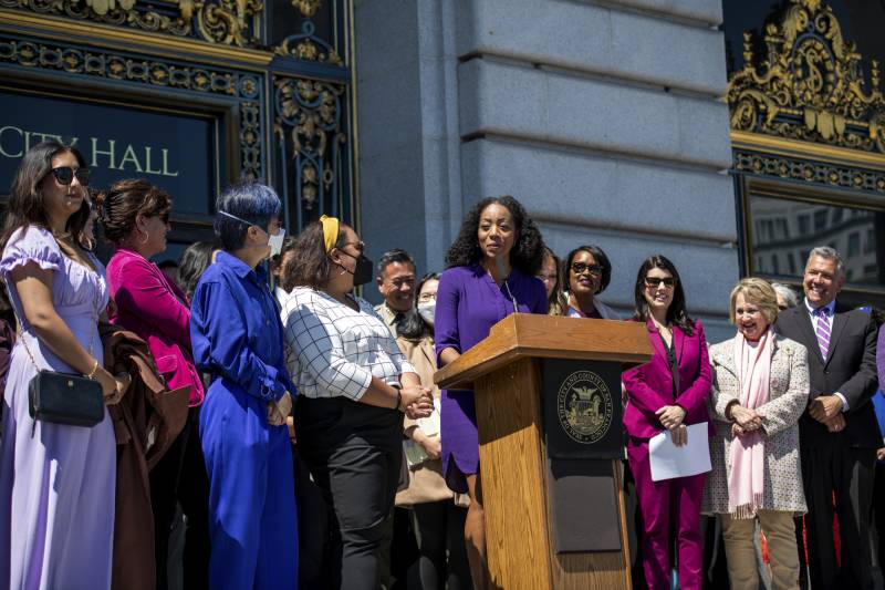 A person in a purple dress speaks at a podium in front of a group of people assembled in front of an ornate building.
