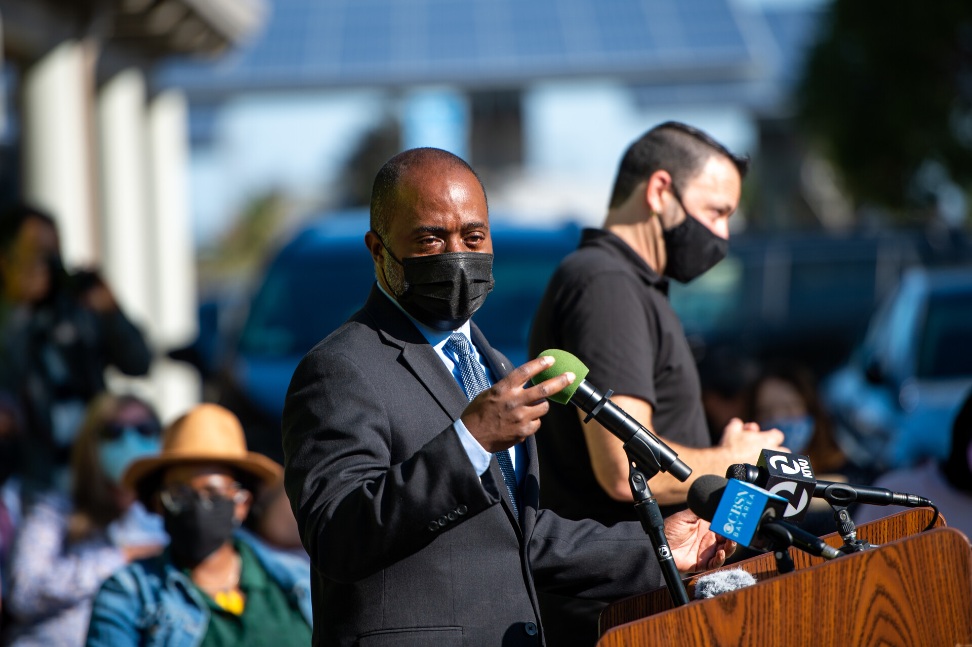 California Superintendent Tony Thurmond is pictured speaking from a wooden podium. He has a business suit and black face mask on. It's daytime.