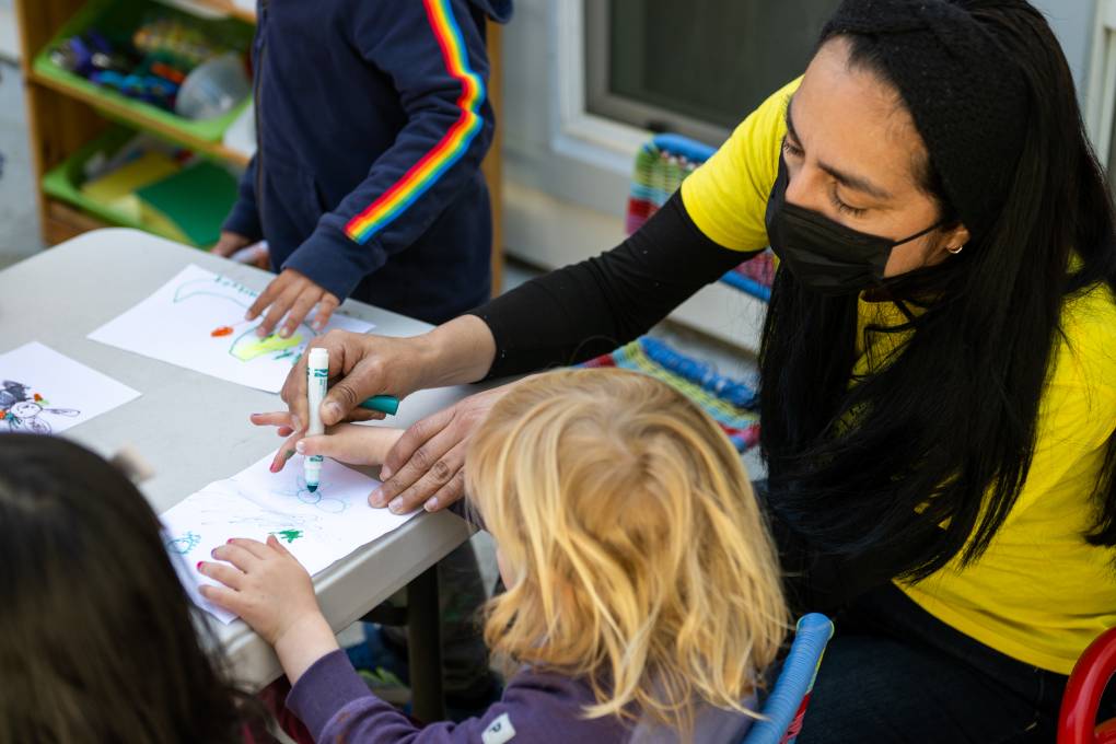 A woman wearing a yellow shirt and black masks leans over a table with small children to draw on a piece of paper in front of a blonde haired child.
