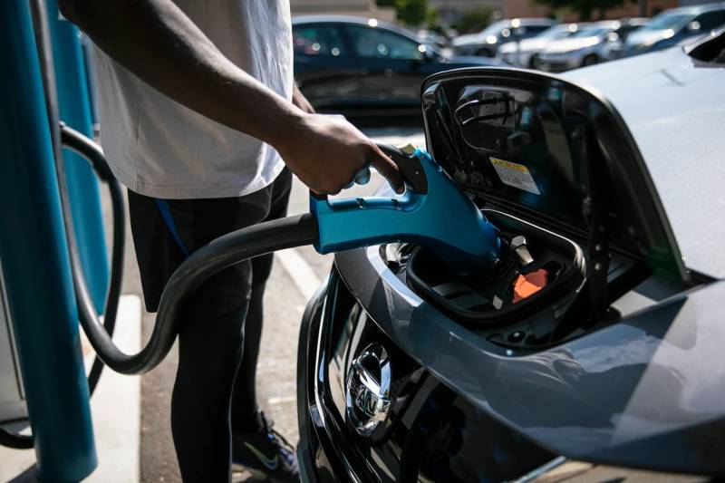 An electric car is charging as a man's arm holds the charger at a charging station.