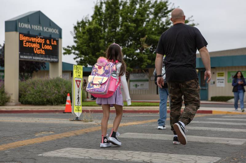 A small girl wearing a purple skirt and pink backpack walks with a bald man wearing a black shirt and camouflage pants in a crosswalk.