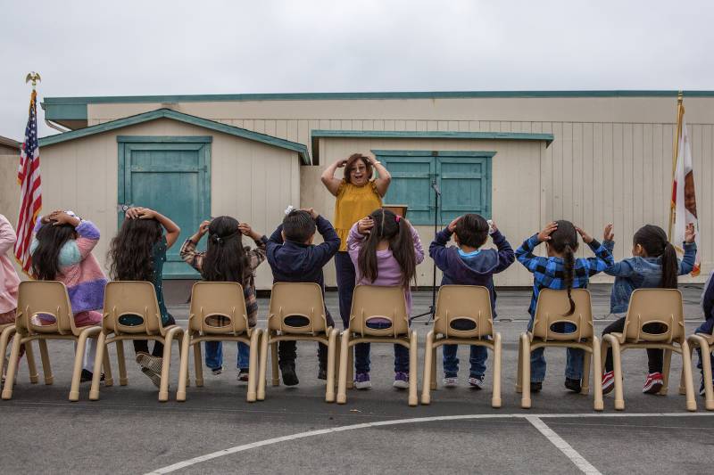 Eight children sit in chairs on a playground with their hands on their heads facing a woman who is doing the same thing while standing.