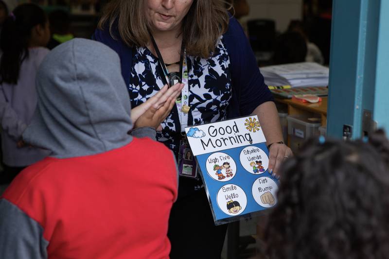 A woman wearing a shirt with blue and white designs holds a book that reads "Good Morning" with illustrations in front of children.