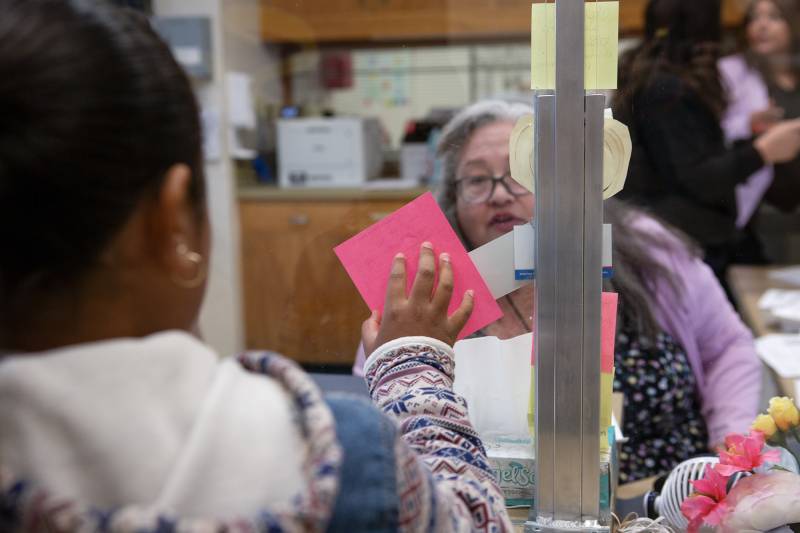 A young woman holds a pink card against a glass window as a woman wearing glasses looks at her.