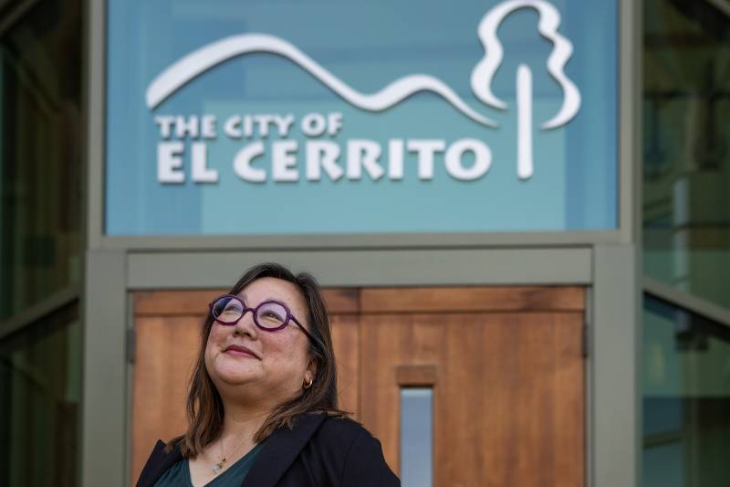 A woman wearing glasses stands below a sign that reads "The City of El Cerrito."