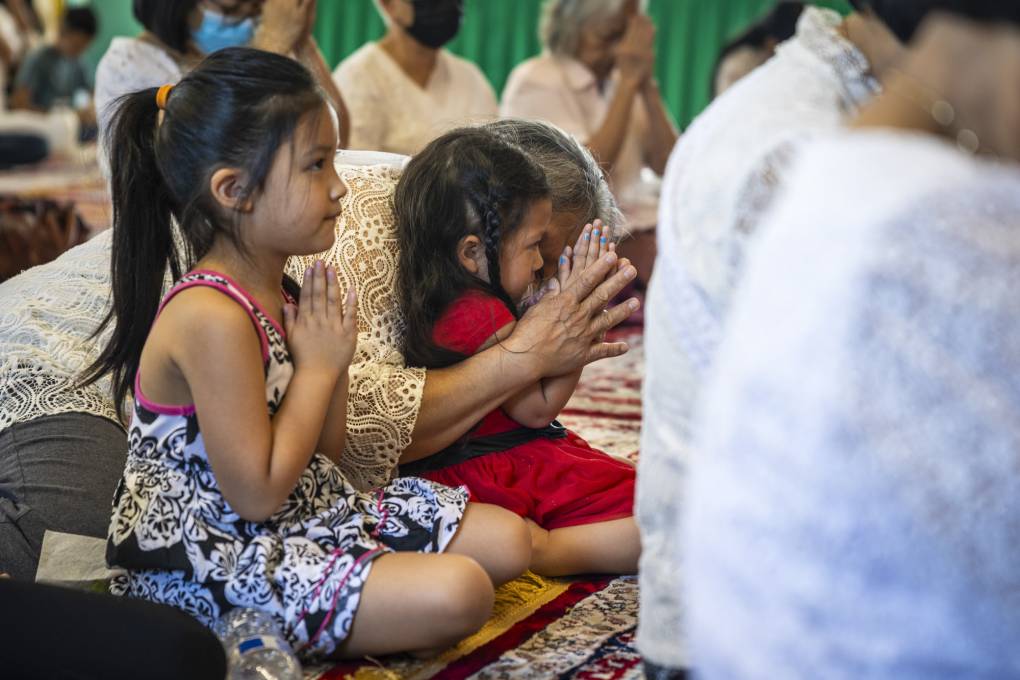 Two young children hold their hands together in prayer, the smaller of which is helped by an elderly person in doing so.
