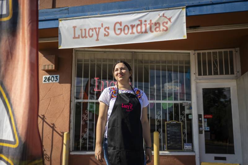 A woman wearing a white shirt with an apron that says "Lucy's" stands in front of building that has a sign that reads "Lucy's Gorditas."
