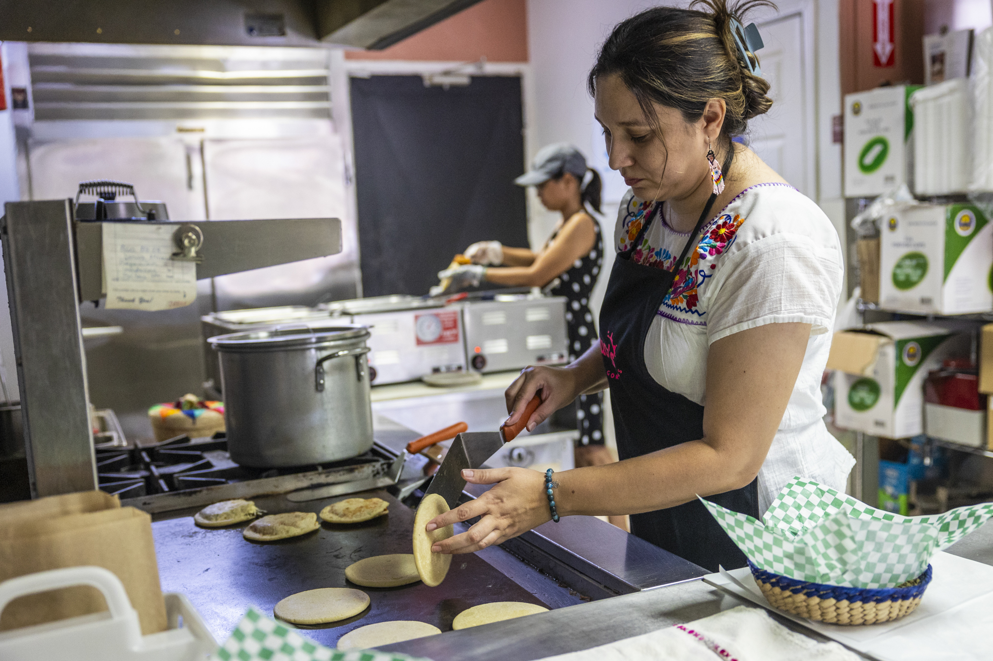 Two women are seen in a kitchen. The woman wearing an apron and closest to the camera turns over a gordita with her hand and utensil.