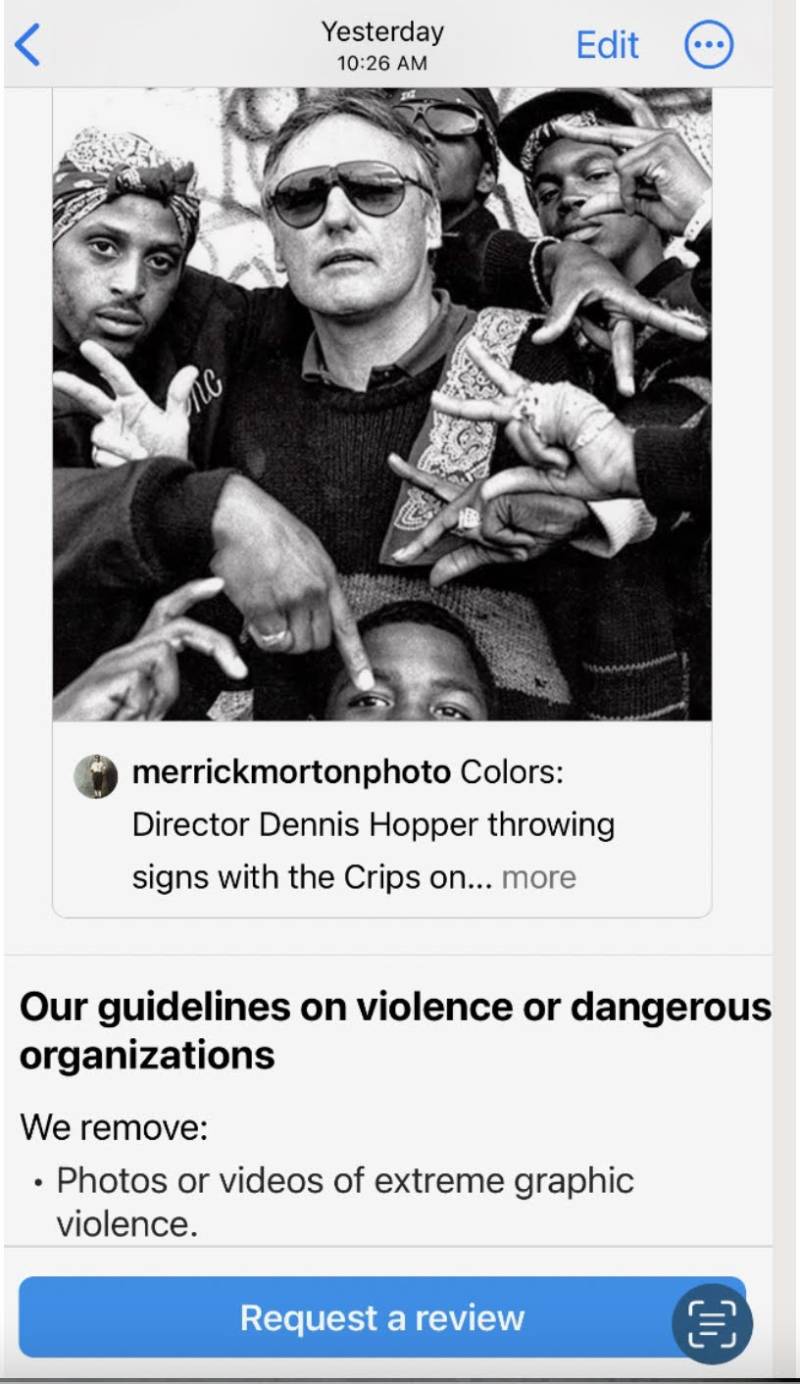 A screen shot of a photo of a man wearing sunglasses surrounded by other people making signs with their hands.
