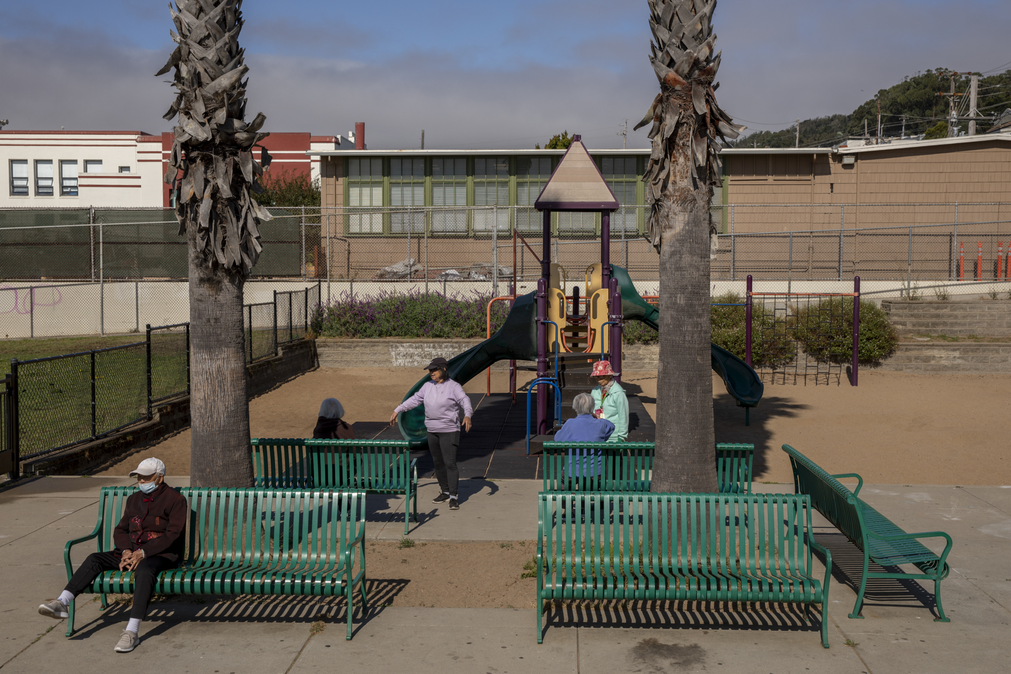 Five people congregate around park benches in front of a jungle gym in an outdoor park.
