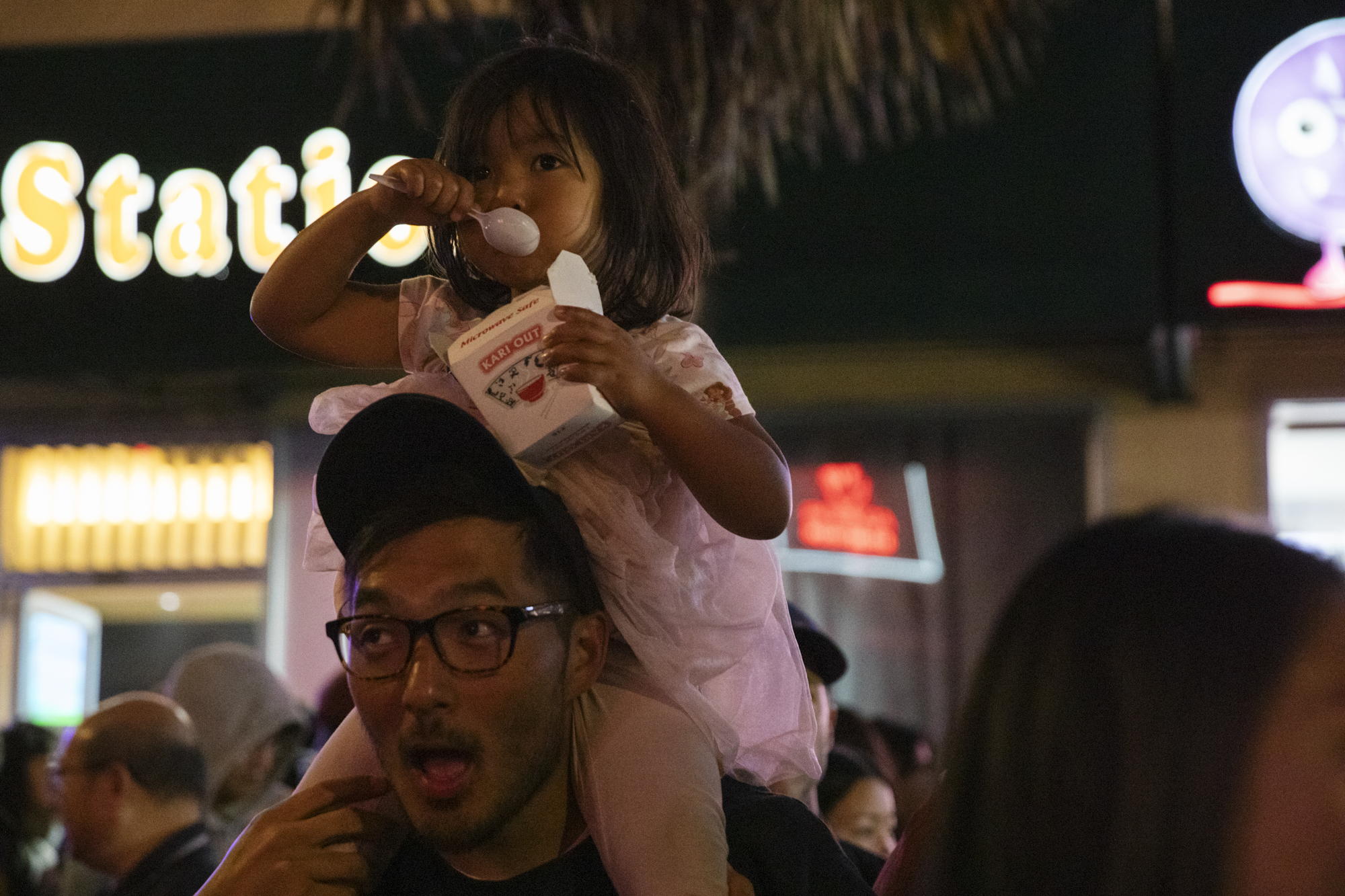 A child eats from a takeout container while sitting on their parent's shoulders.