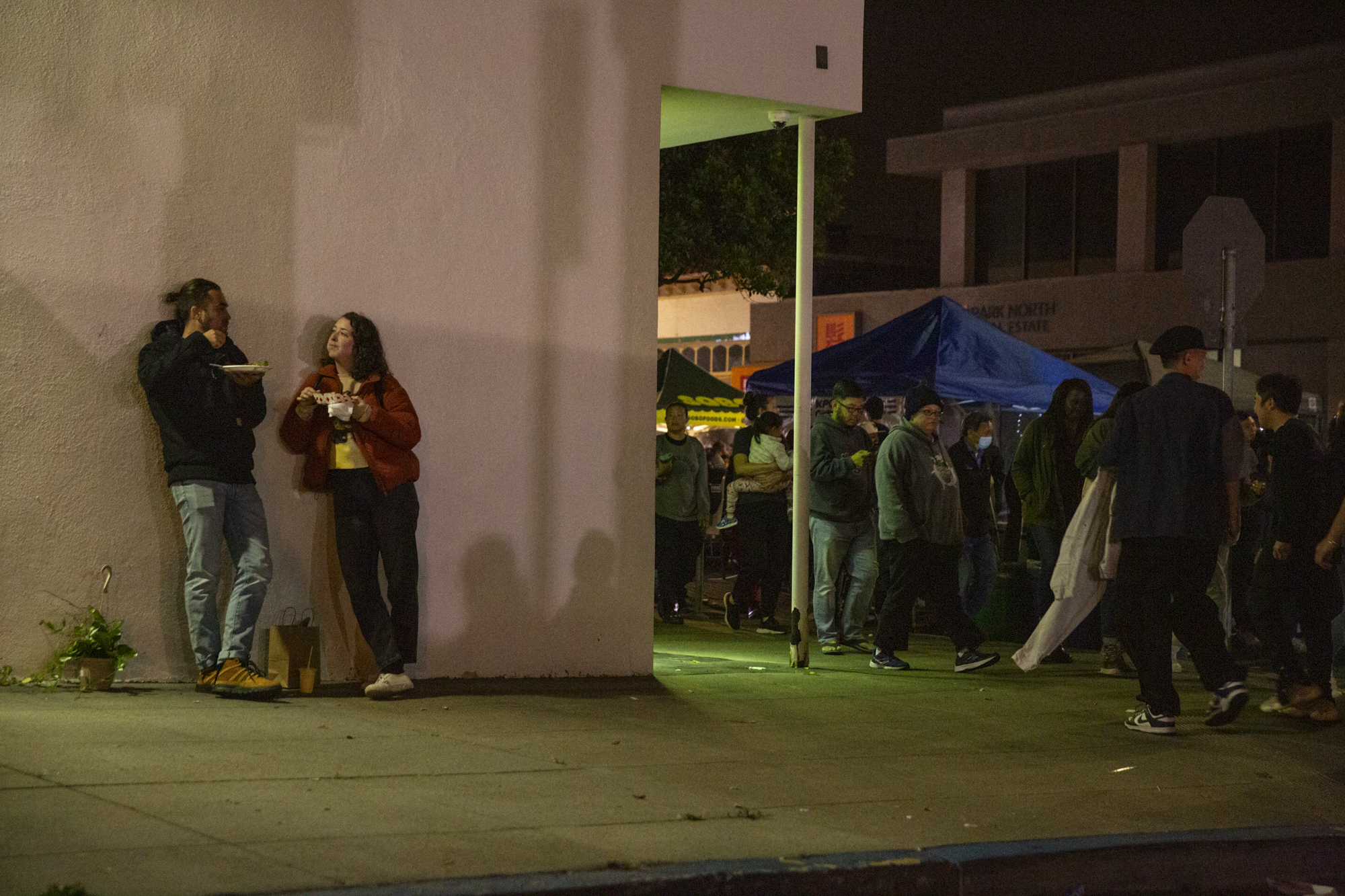 Two people lean on a wall eating while a crowd walks by.