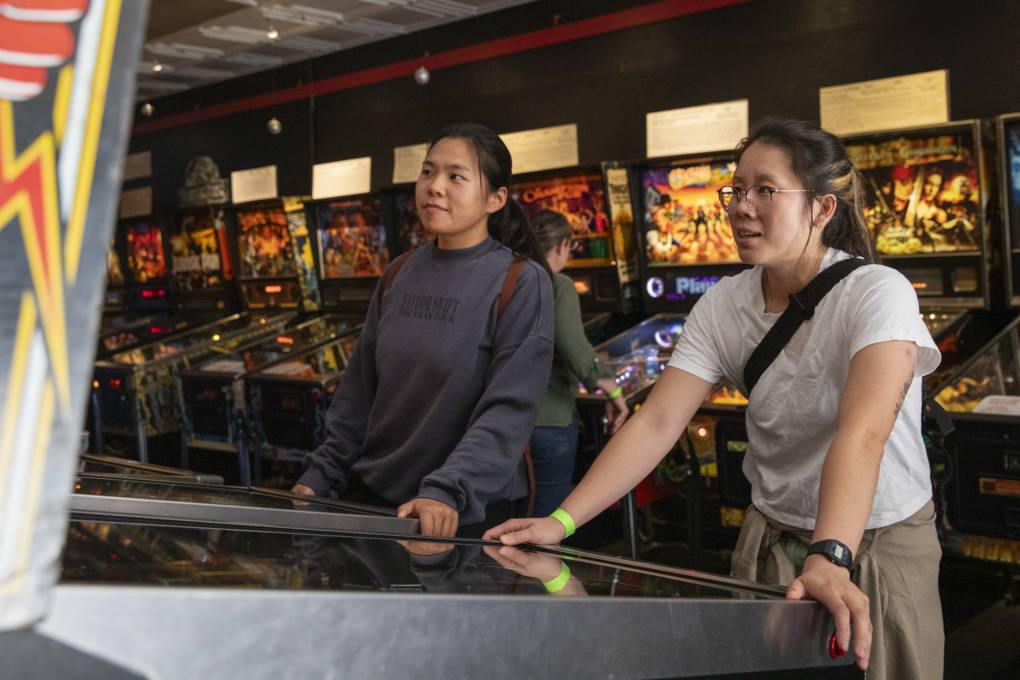 Two people stand in front of pinball machines looking intently at one of them.