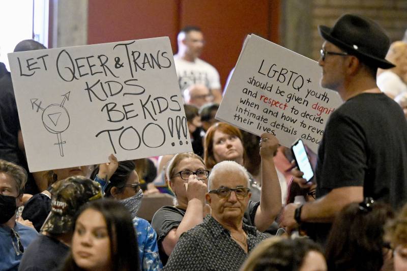 A crowd of people in an indoor setting including some holding signs, one of which reads "Let Queer & Trans Kids Be Kids Too!"