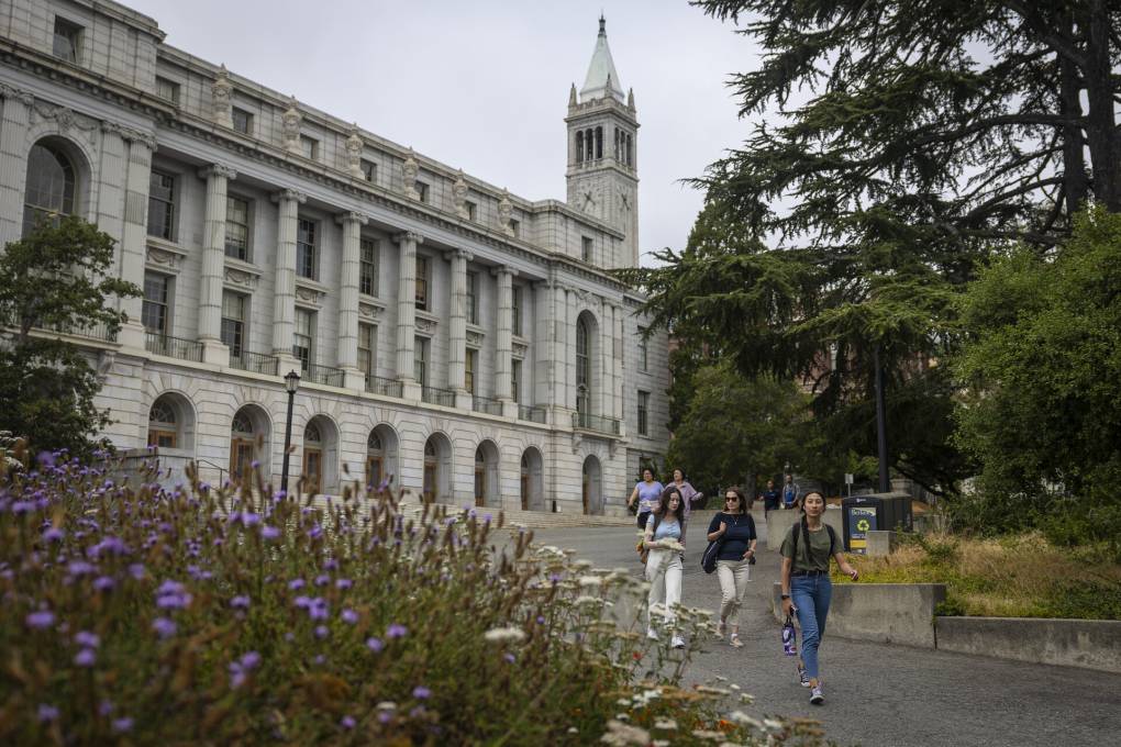 A college campus is pictured with a large building with a pointed clock tower. Students with backpacks are seen walking down a cement pathway on a gray afternoon.