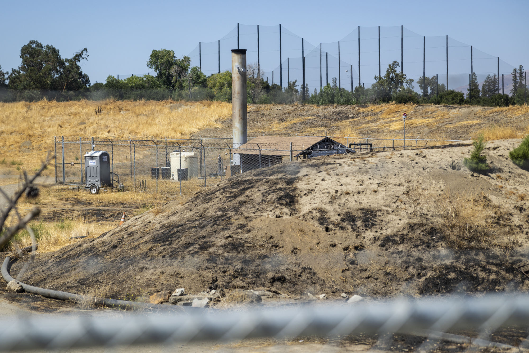 A small structure with a large smokestack is seen from behind a chainlink fence in the center of a large field.