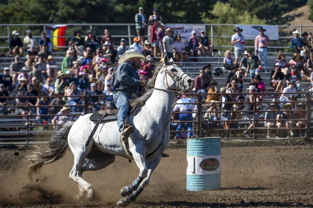 A person wearing cowboy attire rides a horse on a dirt field with people watching from stands.