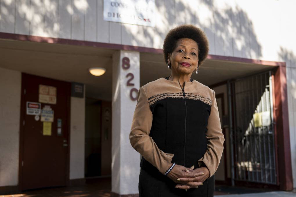 An older African American woman stands in front of a residential building, looking at the camera.