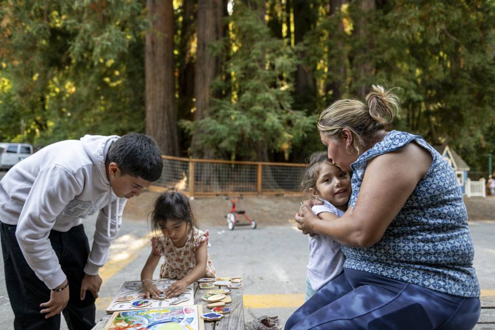 An adult hugs and kisses a child while two other young people do a puzzle in an outdoor setting.