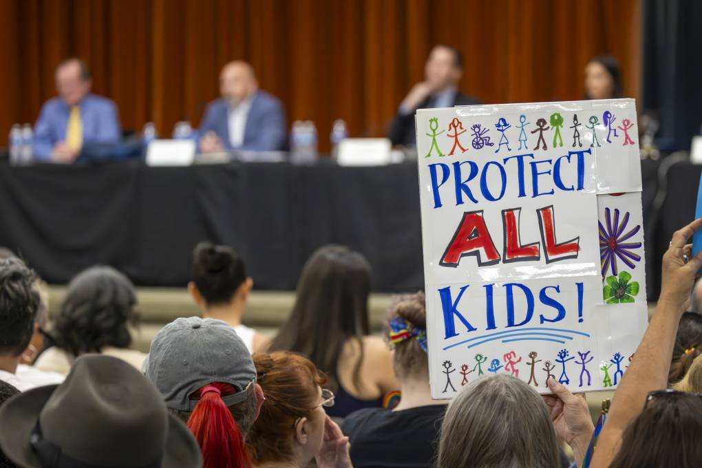 A person holds a sign above their head that reads "protect all kids" in a crowded indoor space.