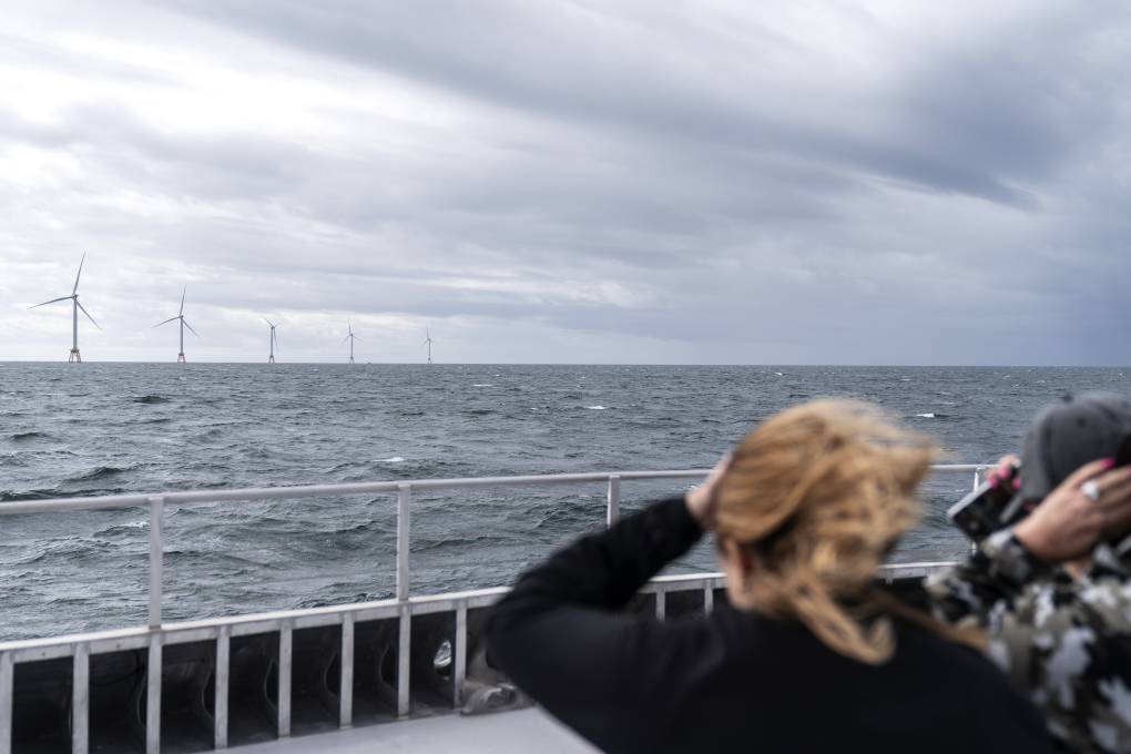 People on a windy pier look out into the wavy ocean and see wind turbines in the distance poking out among the water. The sky is gray and cloudy. One blond woman's hair is blowing in the wind.