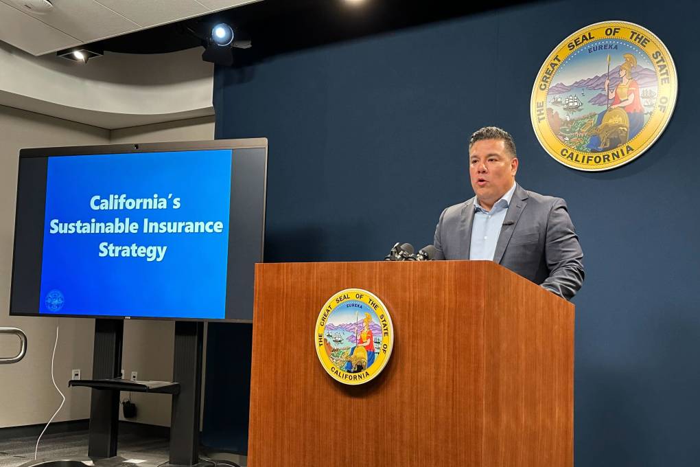 a man in a suit stands at a lectern in front of a sign that says 'California's sustainable insurance strategy'