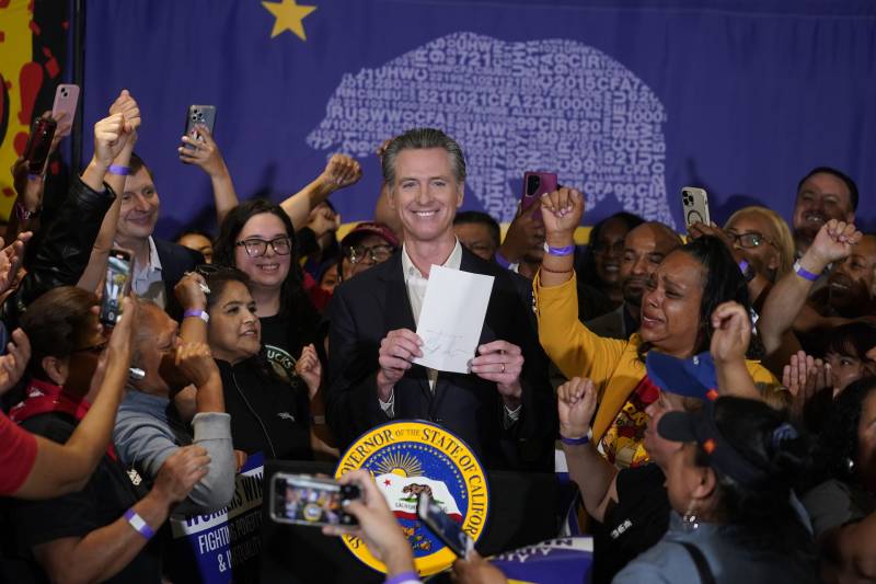 A white man wearing a suit holds a piece of paper at a podium and is surrounded by people cheering and clapping.