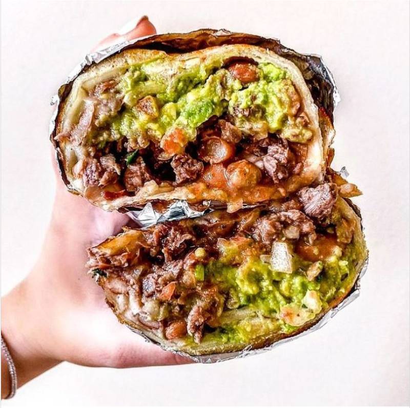 Hand holding a large burrito stuffed with filling and wrapped in foil, cut in half.