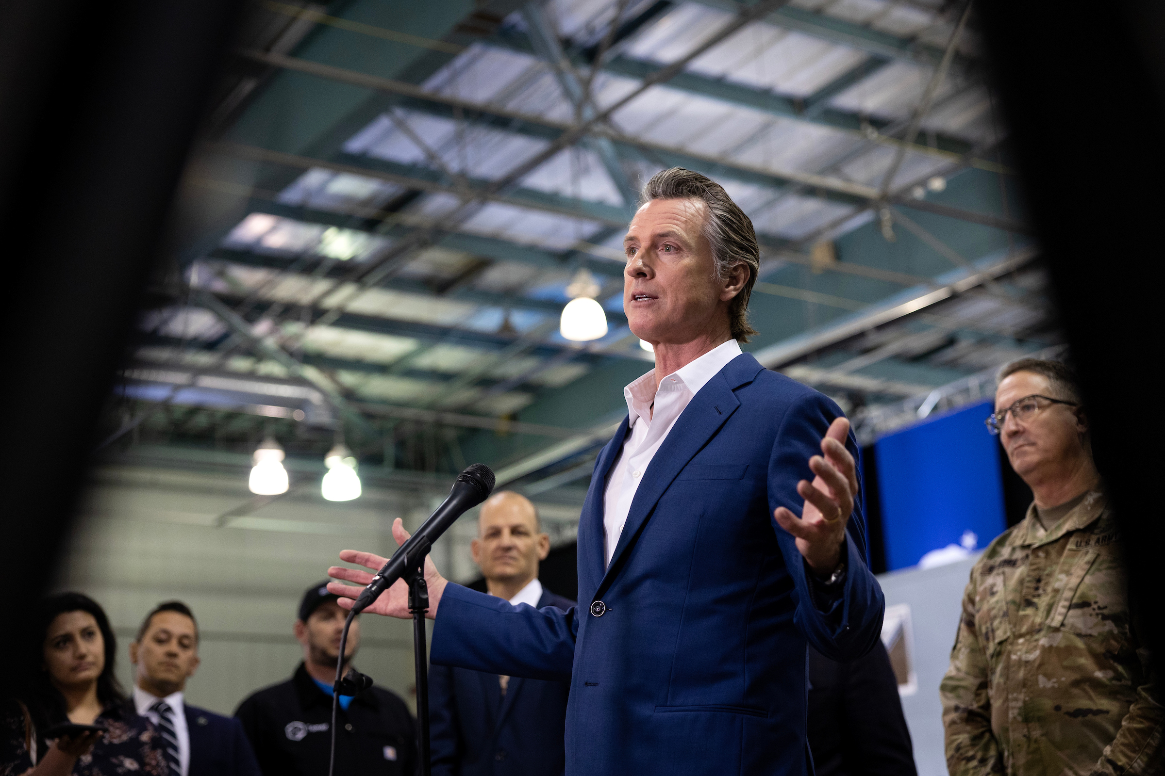 Gov. Gavin Newsom is pictured with his hands out as he speak to many folks in a warehouse in front of a microphone.