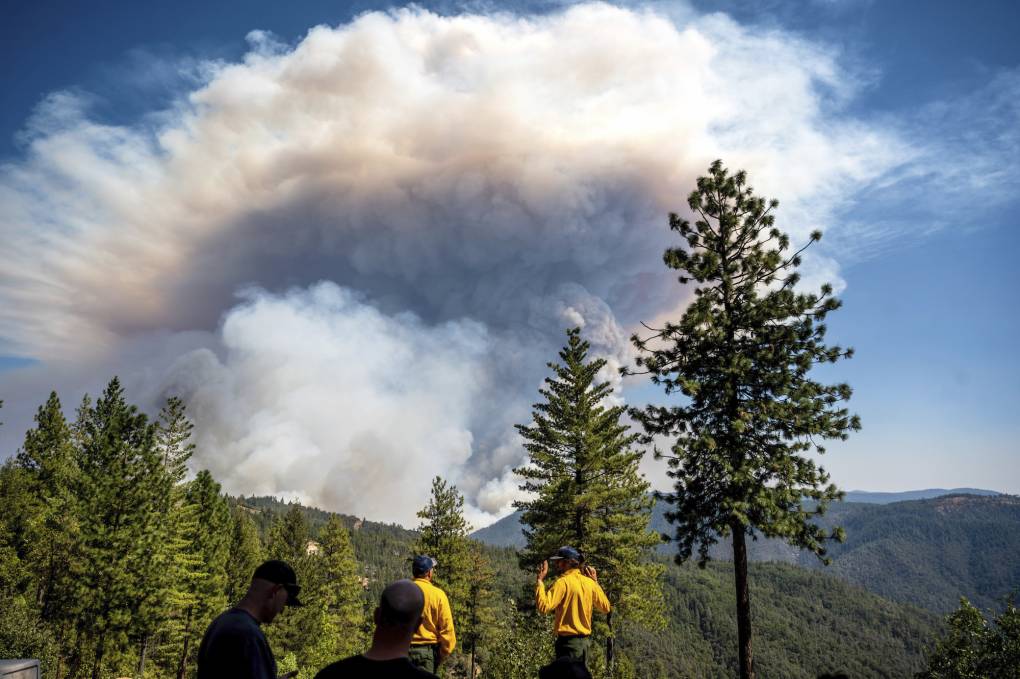 A gigantic, gray plume of wildfire smoke is seen in the distance. Green, rolling mountains with lush trees surround it. Men in yellow uniforms and baseball caps observe the plume from a distance.