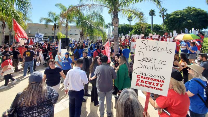 A protest is happening in this photo with many college students of all ages holding signs that say, "Students Deserve Smaller Classrooms. California Faculty Association."
