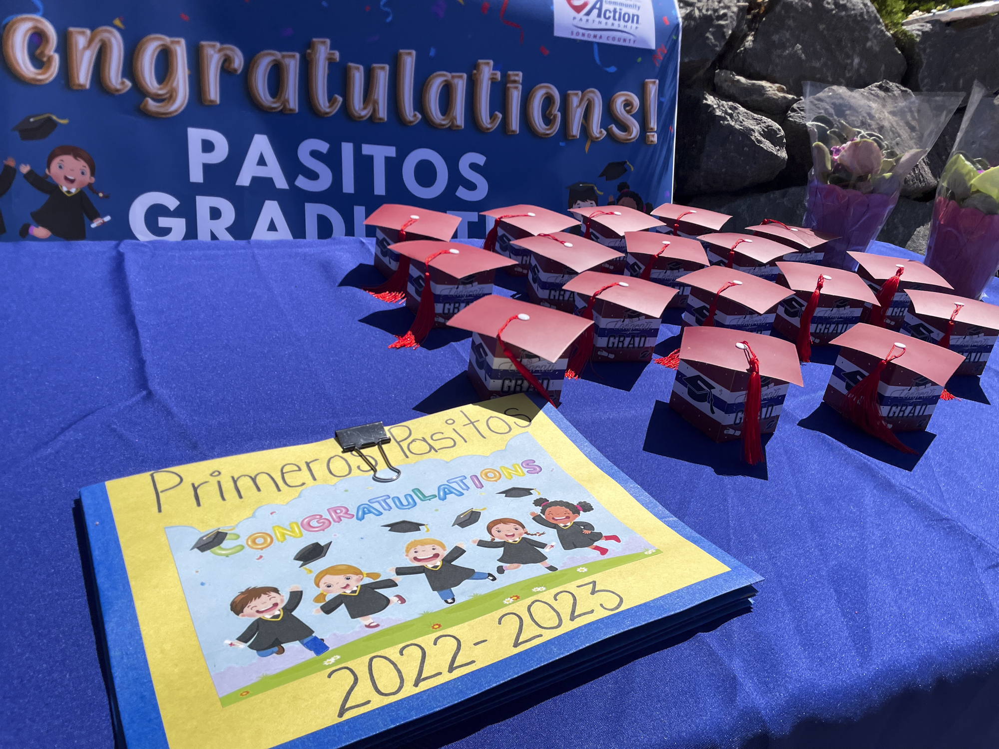 A table top with with a blue table cloth and cardboard graduation caps and paperwork reading "Primeros Pasitos: Congratulations."