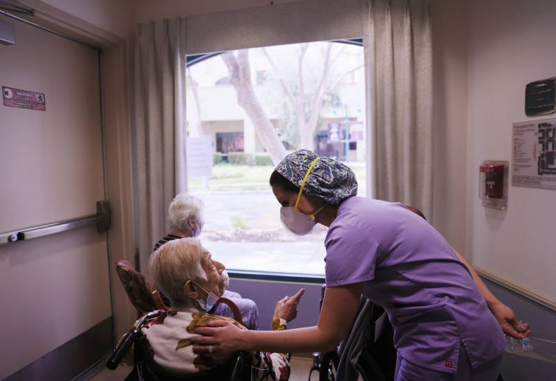 A nurse wearing a mask and hair net addresses a patient in a wheelchair near a window.