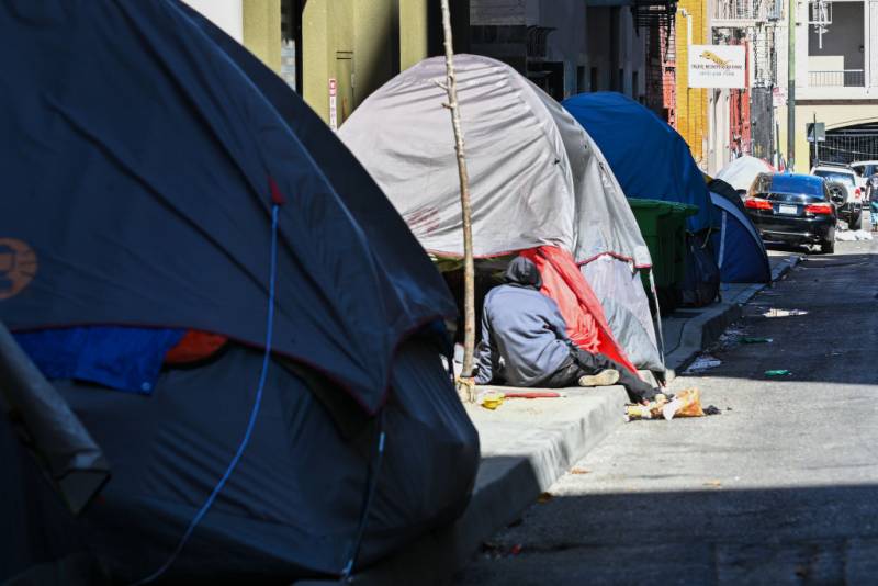 Three tents line a sidewalk, with a person - back to camera - sitting on the curb in front of one of the tents.
