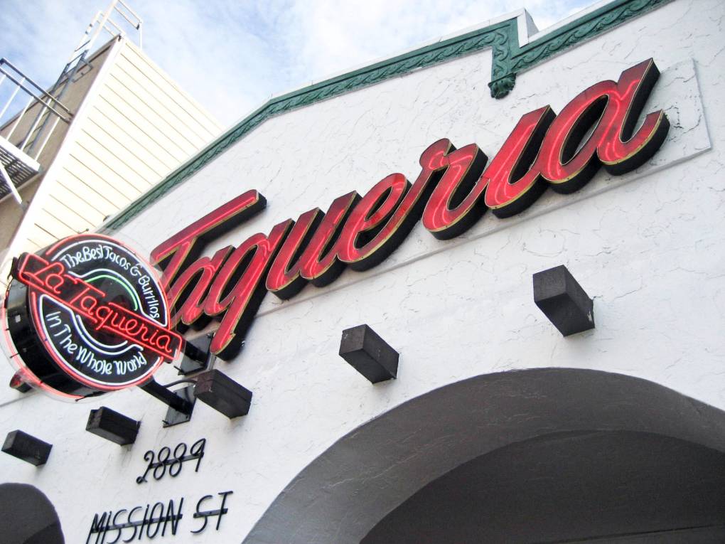 Facade of a building in white stucco. A large red sign reads "La Taqueria," and a smaller neon sign says "La Taqueria. The best tacos and burritos in the whole world."
