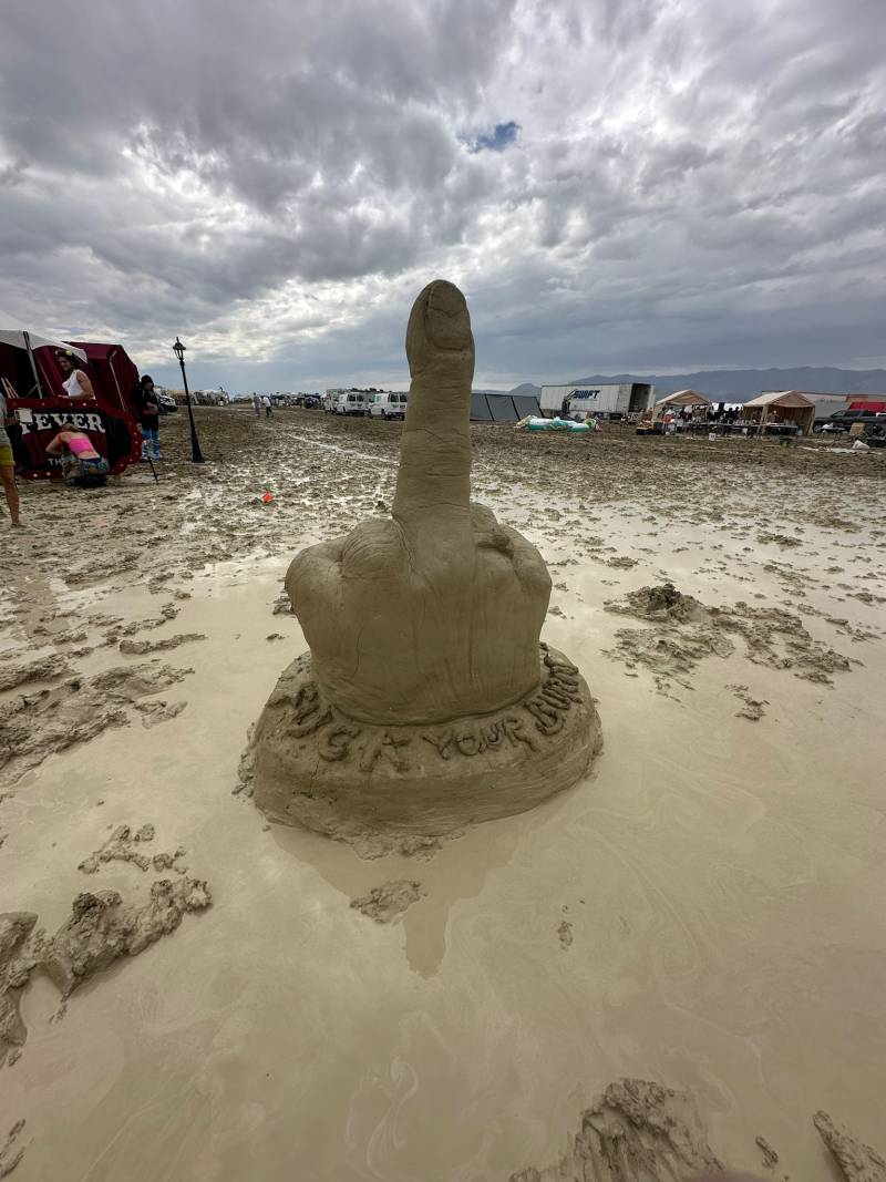 A middle finger made out of mud sits in a muddy scene with clouds in the background. 