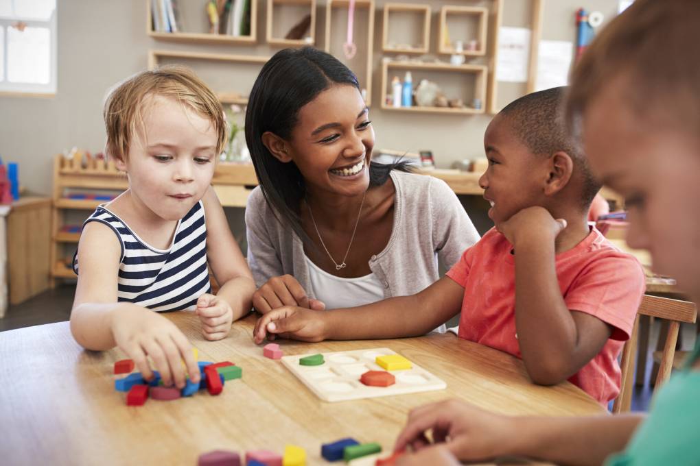 A young Black woman teacher sits at a classroom table smiling at kindergarten-aged children of various races