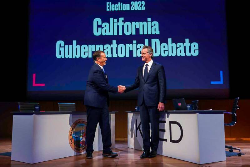 Two men wearing business suits shake hands on stage.