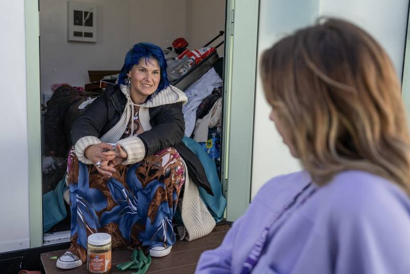 A woman with blue hair a jacket and lots of clothing behind her sits down while a woman wearing purple is in the foreground.