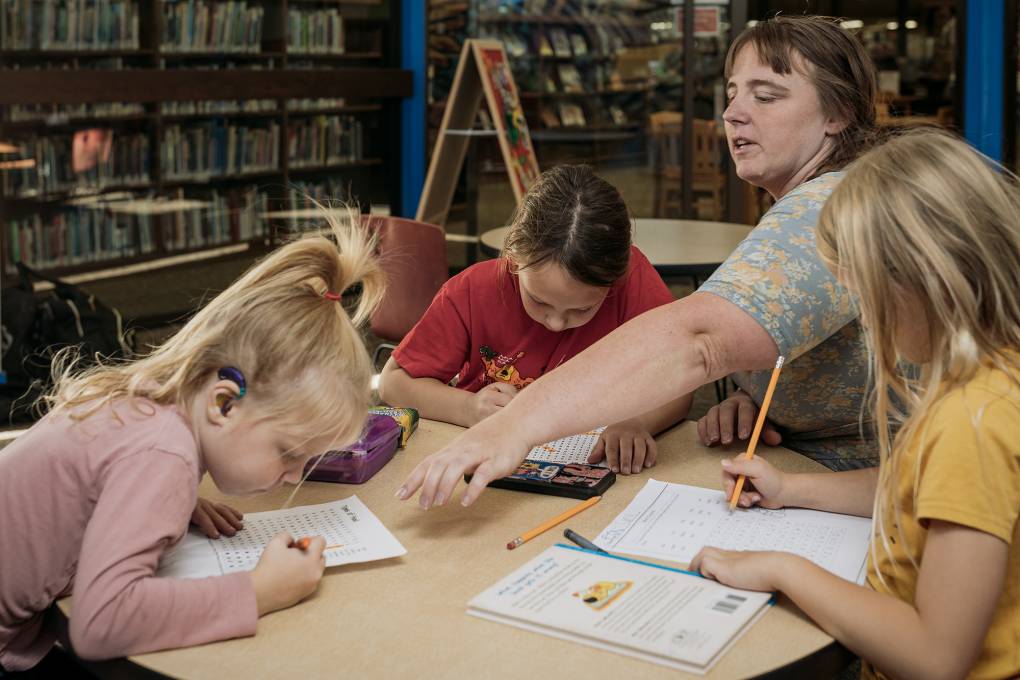 A white woman on the right points at a piece of paper while three young white girls sit around a desk holding pencils on sheets of paper.