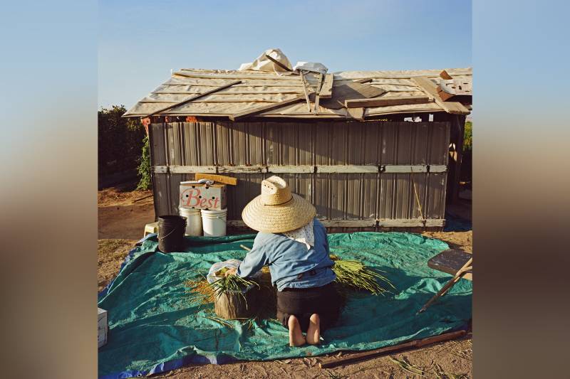 A person kneels outside of a small hut on a blue tarp as they work.