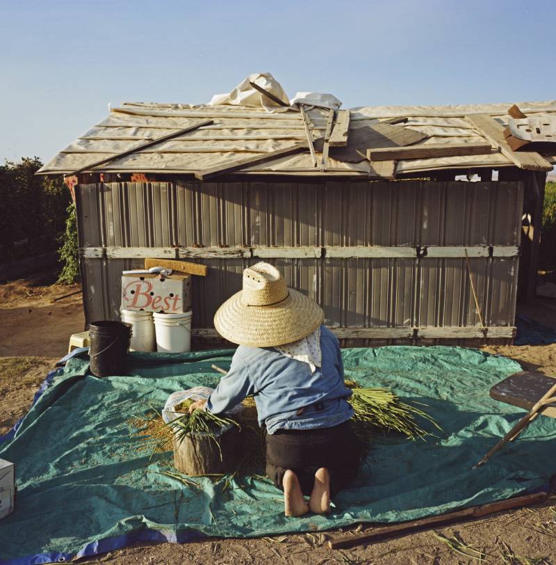 A person kneels outside of a small hut on a blue tarp as they work.